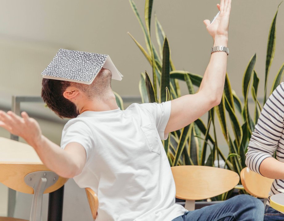 Person leaned back in chair, hands raised to the sky in frustration, a notebook balanced on their face.